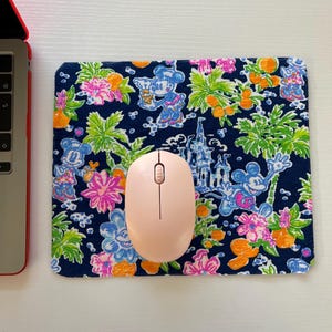 May include: A navy blue mousepad featuring a colorful floral and Mickey Mouse pattern. A light pink wireless mouse sits on the pad. The design includes pink, orange, and green floral elements.