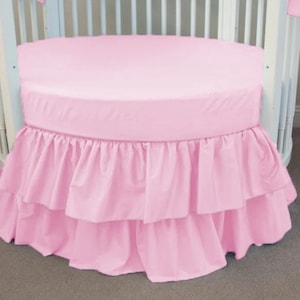 May include: A pink round crib skirt with a ruffled bottom. The skirt has two layers of ruffles.