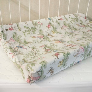 May include: A white changing pad cover with a green, pink, and blue floral pattern featuring people and trees.