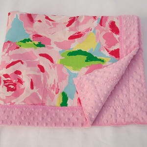 May include: A baby blanket with a floral pattern in pink, red, green, and blue. The blanket has a soft, pink, textured backing and a pink border. The blanket is folded over, showing the textured backing.