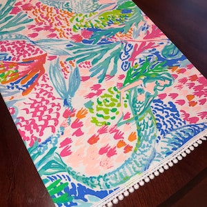 May include: A white table runner with a colorful floral pattern. The pattern features pink, blue, green, and orange flowers and leaves. The runner has a white pom-pom trim.