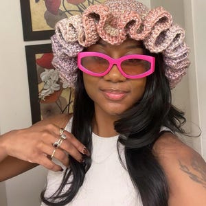 May include: A woman wearing a pink and purple crocheted hat with a ruffled design. She is also wearing pink sunglasses.