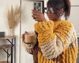 May include: A person wearing a yellow, brown, and beige chunky knit sweater with a cup of coffee in their hand. They are wearing black framed glasses and have their hair pulled back in a ponytail.