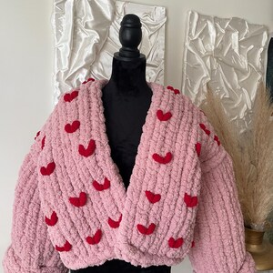 May include: A pink knitted cardigan with red heart embellishments. The cardigan has a V-neck and long sleeves.