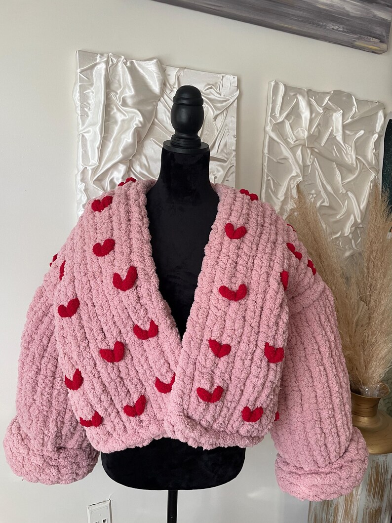 May include: A pink knitted cardigan with red heart embellishments. The cardigan has a V-neck and long sleeves.