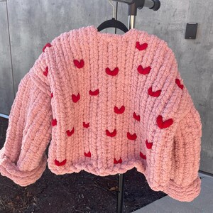 May include: A pink knitted sweater with red heart embellishments. The sweater has a relaxed fit and a cropped length.