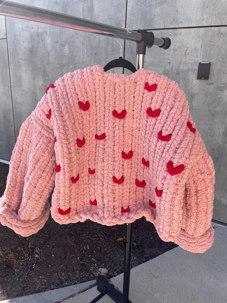 May include: A pink knitted sweater with red heart embellishments. The sweater has a relaxed fit and a cropped length.