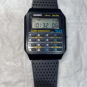 May include: Black Casio CA-50 calculator watch with a black perforated band. The digital display shows the time as 10:32:06 AM. The watch has multiple buttons for alarm and chronograph functions. The watch is a retro style.