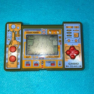 May include: A vintage Casio CG-440 handheld game in olive green and blue. The screen displays a map, with red buttons for fire and control. The text "RADAR POWER" and "GAMER'S RANK" are visible on the device.