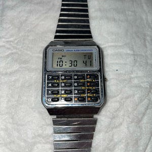 May include: A silver Casio digital watch with a calculator keypad and a segmented metal band. The watch face displays the time as 10:30 PM and the day as Tuesday. The watch has the text "CASIO Lithium ALARM CHRONOGRAPH".