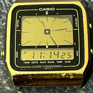 May include: A vintage Casio digital watch with a gold-tone case and a black face. The watch displays the time as 11:14:25. Features include time, date, alarm, and chronograph functions. The watch has a rectangular shape.