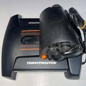 May include: Black Thrustmaster joystick and base. The joystick has a textured grip and an orange button. The base has orange markings and the Thrustmaster logo. A black cable with a yellow band is attached.
