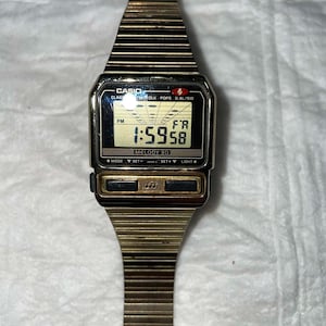 May include: A vintage Casio digital watch with a gold-tone metal band and case. The watch face displays the time as 1:59:58 PM. The watch has several function buttons and the text "MELODY 30" is visible.