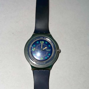 May include: A Swatch watch with a dark blue band and a round face. The watch face is blue with teal and white accents, including the numbers 12, 3, 6, and 9. The watch hands are silver.