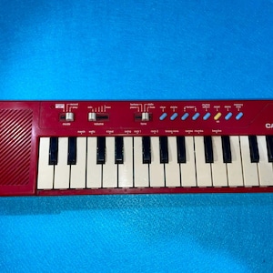 May include: A red Casio PT-10 keyboard with black and white keys. The keyboard has various control buttons and sliders, including volume and mode. The text "CASIO PT-10" is printed on the right side of the keyboard.