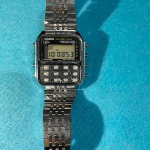 May include: A silver-toned Casio CA-851 digital watch with a calculator keypad. The watch displays the time as 10:08:53 AM. The watch has a silver link band and a black face with white text. The watch is on a blue surface.