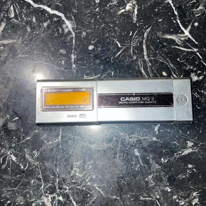May include: A silver Casio MQ-2 Micro Computer Quartz watch with a rectangular LCD screen displaying the time. The watch has buttons for alarm and timer functions. The watch is on a black and white marble surface.