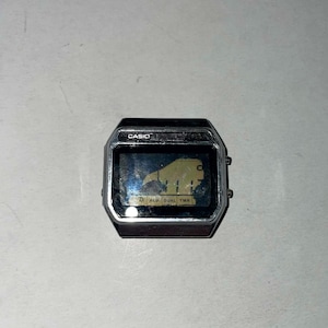 Puede incluir: Un reloj digital Casio vintage con caja plateada. La esfera del reloj muestra la hora digital y un gráfico. La palabra "CASIO" está impresa encima de la pantalla. El reloj tiene dos botones en el lado derecho.