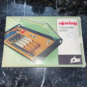 May include: A vintage Rotring Tuschefüller variant pen set in its original box. The box is light green with the Rotring logo and product information. Inside, the set includes several black and silver pens and accessories, nestled in a yellow-orange velvet-lined tray.