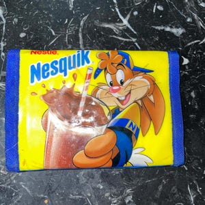 Puede incluir: Una cartera amarilla con borde azul con el logo de Nesquik y un conejo de dibujos animados sosteniendo un vaso de leche con chocolate con una pajita. La cartera tiene una superficie texturizada.