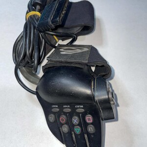 Puede incluir: Mando de juegos negro con correas para la muñeca. El mando tiene botones etiquetados como 'Reset', 'Start', 'Select' y los símbolos de PlayStation. Un cable y un conector negros están adjuntos.