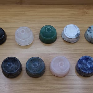 May include: A collection of ten gemstone bottle stoppers. The stoppers are round with a flat top and a slightly tapered base. The colours vary, including black, clear, green, white, and blue. They are arranged on a light brown wooden surface. Each stopper is approximately 3 cm in diameter.
