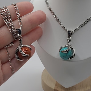 May include: Two silver necklaces with claw pendants holding gemstone spheres. One pendant has a reddish-brown sphere, the other a turquoise sphere. The necklaces are displayed on a white mannequin and held by a hand.