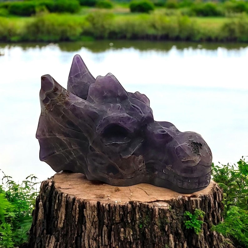 A carved amethyst dragon head sculpture. The sculpture is a deep purple color and has intricate details. The sculpture is sitting on a tree stump.