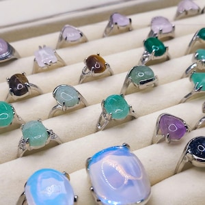 May include: A collection of rings featuring a variety of gemstone settings, including green, pink, purple, and brown stones. The rings are presented in a white box with individual compartments.