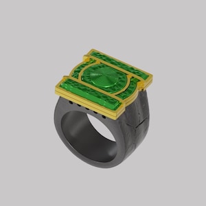 Op de afbeelding: Een ring met een donkergrijze band met een getextureerd oppervlak en een rechthoekig bovendeel. Het bovendeel is goudkleurig met een felgroen, cirkelvormig, geribbeld embleem in het midden en groene, getextureerde zijpanelen voor een geometrisch ontwerp.
