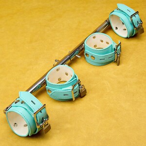 Op de afbeelding: Een set van vier turquoise leren handboeien met zilveren gespen en hangsloten, bevestigd aan een zilveren metalen staaf. De handboeien zijn in een rij gerangschikt, met een gele achtergrond.