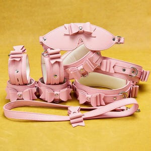 Op de afbeelding: Een verzameling roze leren boeien, waaronder handboeien, een blinddoek en een halsband, allemaal versierd met decoratieve strikken. De set bevat ook een bijpassende riem. Op een gele achtergrond.