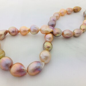 May include: A strand of iridescent baroque pearls in shades of pink, peach, and gold. The pearls are strung on a white thread and are arranged in a figure eight shape.