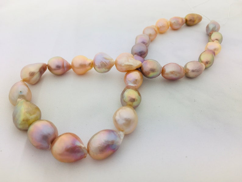 May include: A strand of iridescent baroque pearls in shades of pink, peach, and gold. The pearls are strung on a white thread and are arranged in a figure eight shape.