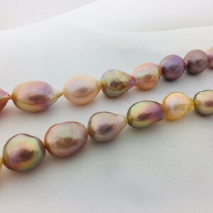 May include: A strand of baroque pearls with a variety of iridescent colors, including pink, peach, and gold. The pearls are strung on a white cord and are arranged in a loose, flowing pattern.
