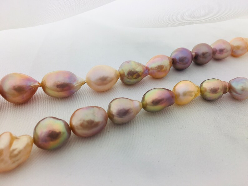 May include: A strand of baroque pearls with a variety of iridescent colors, including pink, peach, and gold. The pearls are strung on a white cord and are arranged in a loose, flowing pattern.