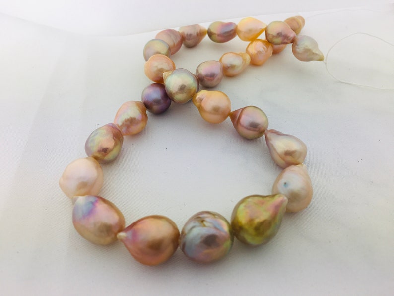 May include: A strand of iridescent baroque pearls in shades of pink, gold, and purple. The pearls are strung on a white thread and are arranged in a circular pattern.