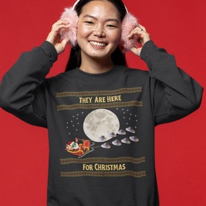May include: A black sweatshirt with a Christmas-themed design featuring a cartoon Santa Claus flying in a sleigh pulled by flying saucers. The text on the sweatshirt reads "They are here for Christmas."