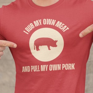 May include: Red t-shirt with a white circle and a pig silhouette. The text on the shirt reads "I rub my own meat and pull my own pork."