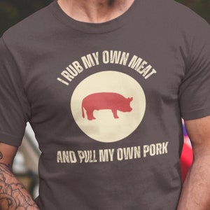 May include: A man wearing a dark gray t-shirt with the text "I rub my own meat and pull my own pork" and a silhouette of a pig in a circle.