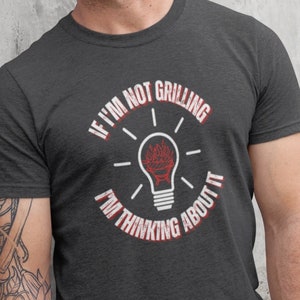 May include: A dark heather grey t-shirt with a white graphic of a lightbulb with flames inside. The text "If I'm not grilling I'm thinking about it" is printed around the lightbulb.