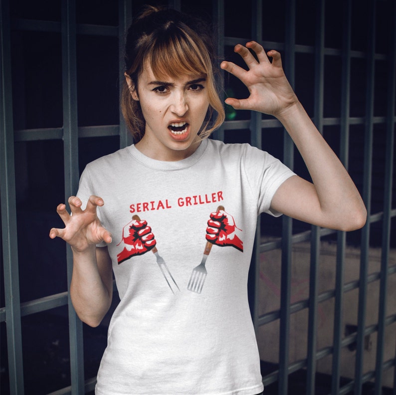 May include: White t-shirt with the text "SERIAL GRILLER" in red letters. Two red hands holding a fork and spatula are printed on the shirt.