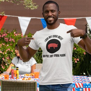 A man standing at a backyard Bbq cookout pointing at his shirt that says I Rub My Own Meat And Pull My Own Pork featuring a graphic of a pig in the middle.