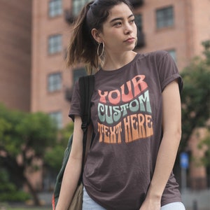 May include: A brown t-shirt with the text "YOUR CUSTOM TEXT HERE" in a retro, colorful font. The text is in shades of orange, green, and blue.