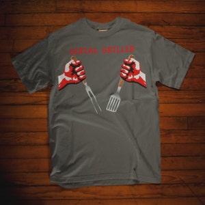 May include: A gray t-shirt with the text "SERIAL GRILLER" in red letters. Two red fists hold a spatula and a fork.