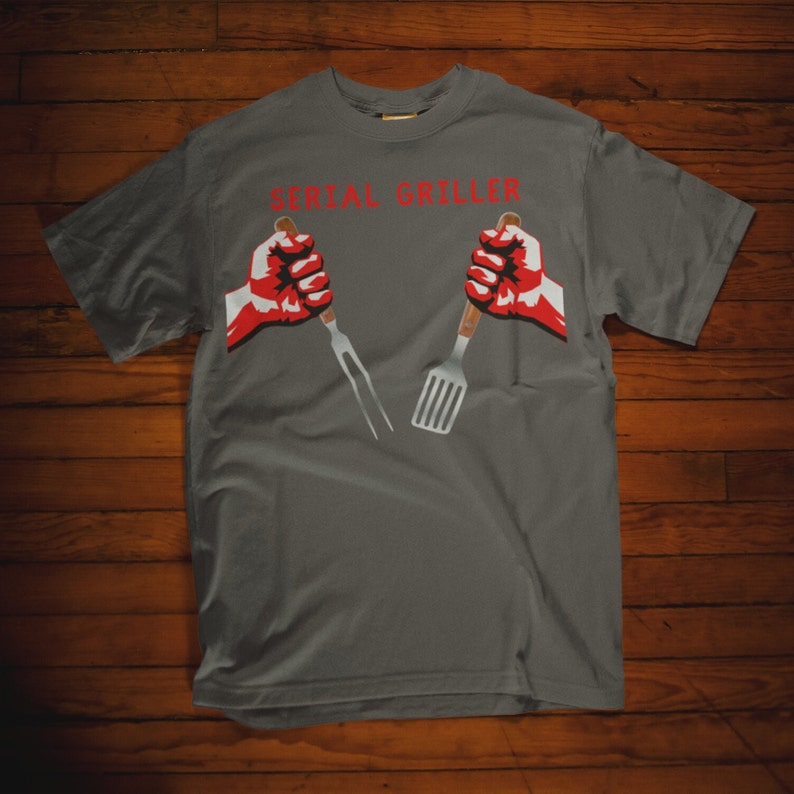 May include: A gray t-shirt with the text "SERIAL GRILLER" in red letters. Two red fists hold a spatula and a fork.