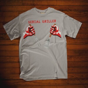 May include: A gray t-shirt with the text "SERIAL GRILLER" printed in black. Two red fists are holding a spatula and a fork.