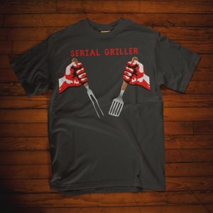 May include: A dark gray t-shirt with the text "SERIAL GRILLER" in white letters. Two red fists are holding a fork and spatula.