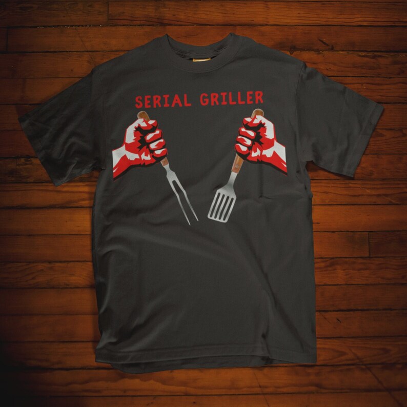 May include: A dark gray t-shirt with the text "SERIAL GRILLER" in white letters. Two red fists are holding a fork and spatula.