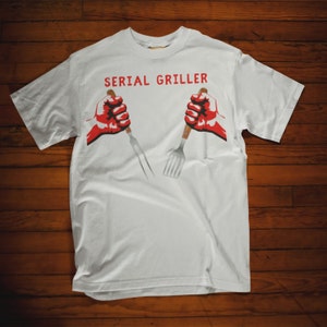 May include: White t-shirt with the text "SERIAL GRILLER" in red letters. Two red fists are holding a fork and spatula.